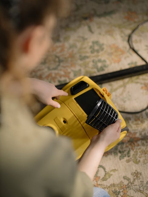 A person holding a yellow portable dehumidifier or air purifier with a black vent, positioned over a worn, patterned carpet with muted tones of beige, green, and red, in a room illuminated by natural 