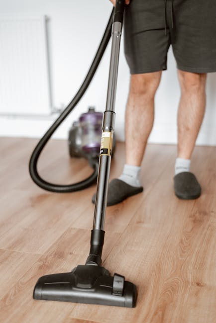 A person holding a yellow portable dehumidifier or air purifier with a black vent, positioned over a worn, patterned carpet with muted tones of beige, green, and red, in a room illuminated by natural 